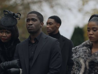 Young man surrounded by a young woman and older woman, dressed in glamourous black outfits with fur, stand outside looking sad. They look like they are at a funeral. 