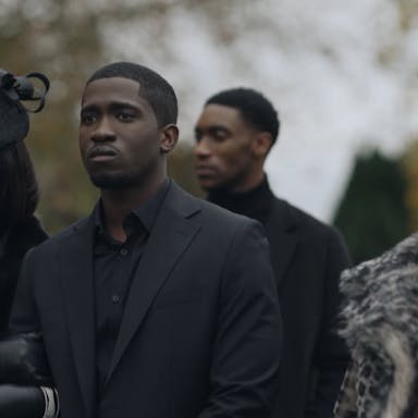 Young man surrounded by a young woman and older woman, dressed in glamourous black outfits with fur, stand outside looking sad. They look like they are at a funeral. 