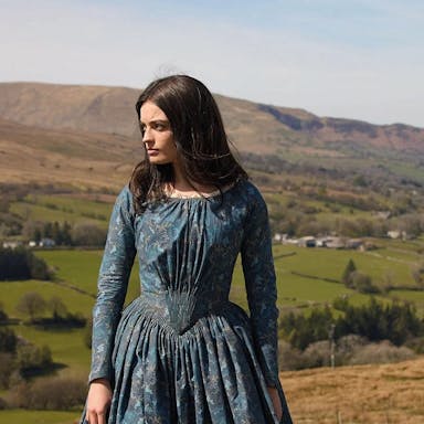 Woman with dark hair in period costume against a countryside backdrop.