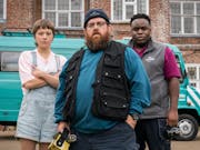 A white woman, white man and Black man stand looking determined in front of a turquoise van