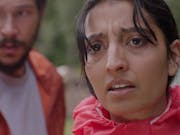 Woman in red raincoat in the foreground and man in orange coat in the background, both look worried.