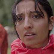 Woman in red raincoat in the foreground and man in orange coat in the background, both look worried.