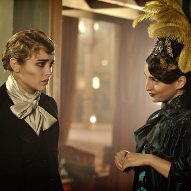 Victorian era, a white woman in male drag is dressed for a performance stood next to a smiling woman with large feathers in her hair