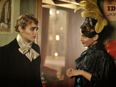 Victorian era, a white woman in male drag is dressed for a performance stood next to a smiling woman with large feathers in her hair