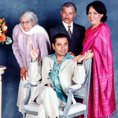 A brown man sits on a chair with two older brown women and an older brown man stand around him with a vase of flowers beside them