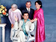 A brown man sits on a chair with two older brown women and an older brown man stand around him with a vase of flowers beside them