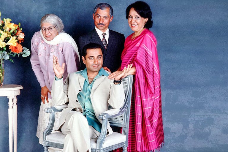 A brown man sits on a chair with two older brown women and an older brown man stand around him with a vase of flowers beside them