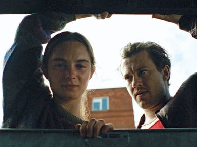 A young girl and an older man, lift the lid and peer through a large industrial waste bin