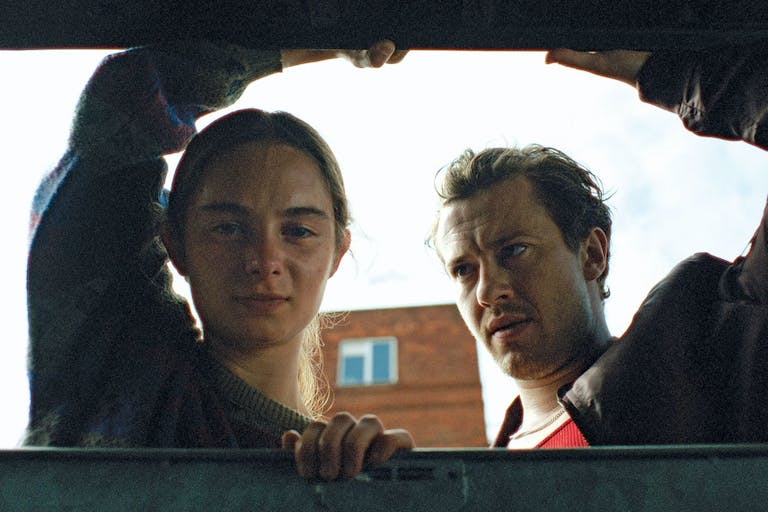 A young girl and an older man, lift the lid and peer through a large industrial waste bin