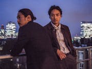 Two Japanese men stand on a high balcony overlooking a cityscape with brightly lit high-rises