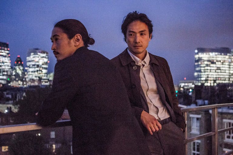 Two Japanese men stand on a high balcony overlooking a cityscape with brightly lit high-rises