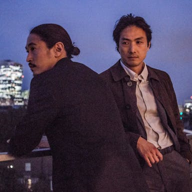 Two Japanese men stand on a high balcony overlooking a cityscape with brightly lit high-rises 