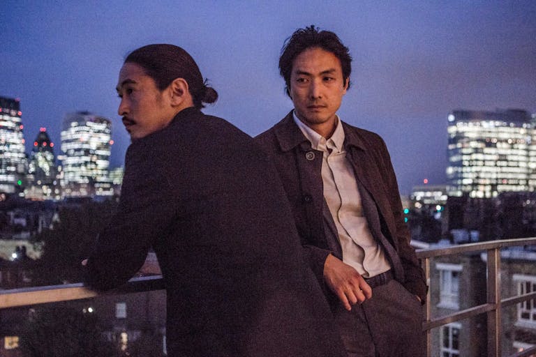 Two Japanese men stand on a high balcony overlooking a cityscape with brightly lit high-rises 