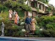 An elderly white man is being helped down stone steps of a beautiful lush green garden with a large house and a few people in the background