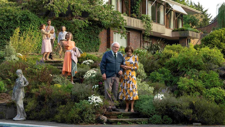 An elderly white man is being helped down stone steps of a beautiful lush green garden with a large house and a few people in the background
