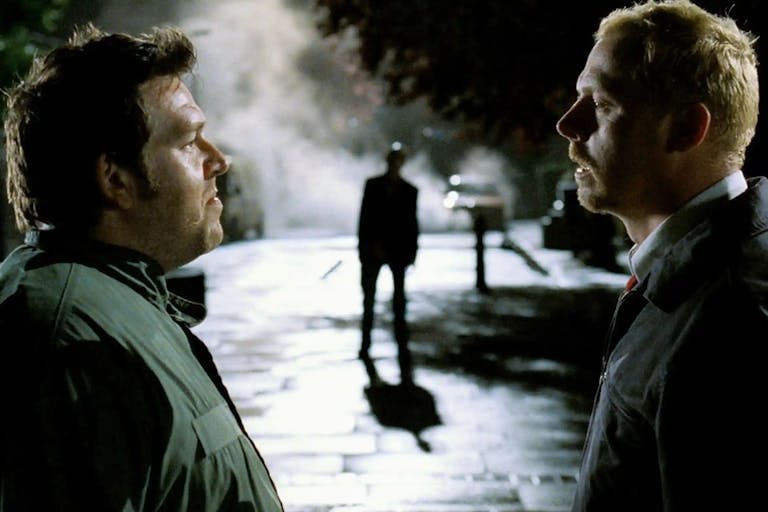 Two white men stand facing each other on the street at night with a ghoulish figure in the background between them