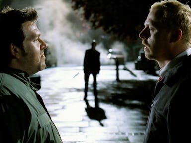 Two white men stand facing each other on the street at night with a ghoulish figure in the background between them