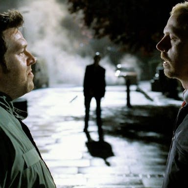 Two white men stand facing each other on the street at night with a ghoulish figure in the background between them