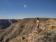 A white woman stands on a dramatic rocky cliff side with bright blue skies in the background