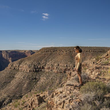 A white woman stands on a dramatic rocky cliff side with bright blue skies in the background