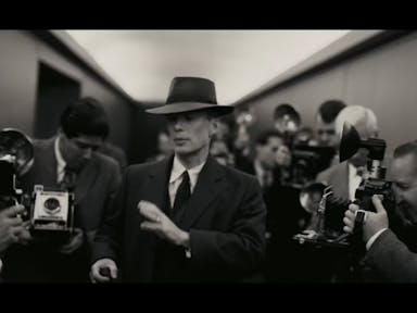 Black and white still of a white man in a sharp suit and hat flocked by photographers with old style flash cameras