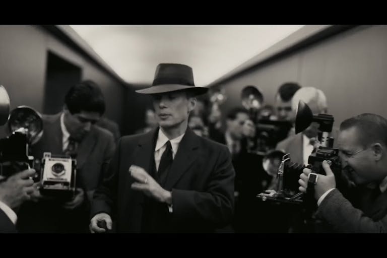Black and white still of a white man in a sharp suit and hat flocked by photographers with old style flash cameras