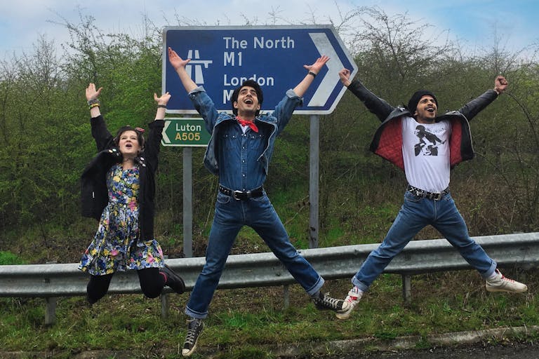 Two young men in denim and a young woman jump in the air with joy, in front of a British motorway road sign.