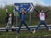 Two young men in denim and a young woman jump in the air with joy, in front of a British motorway road sign.
