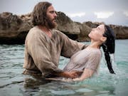A bearded, shaggy haired white man, baptising a white woman with long dark hair in the sea