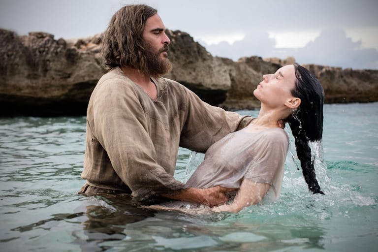 A bearded, shaggy haired white man, baptising a white woman with long dark hair in the sea