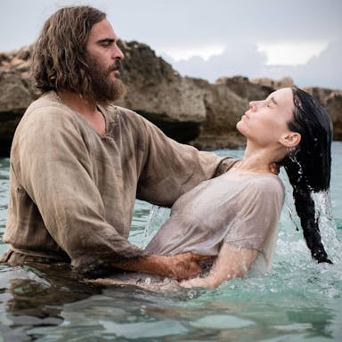 A bearded, shaggy haired white man, baptising a white woman with long dark hair in the sea