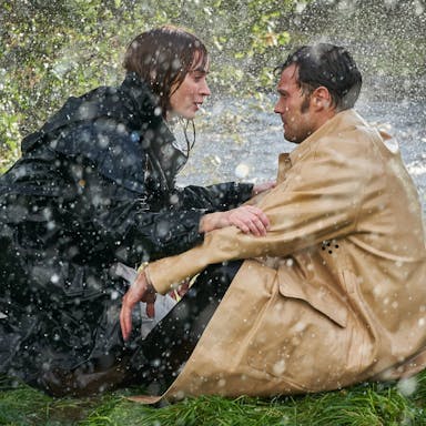 A dark-redheaded white woman and a dark haired white man sit on the edge of a river bank in the rain facing each other, staring intently 