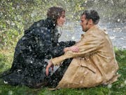 A dark-redheaded white woman and a dark haired white man sit on the edge of a river bank in the rain facing each other, staring intently