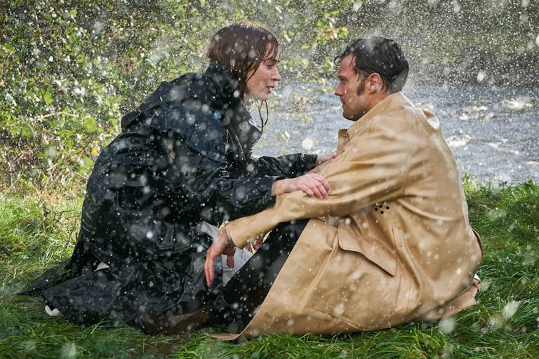 A dark-redheaded white woman and a dark haired white man sit on the edge of a river bank in the rain facing each other, staring intently 