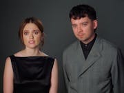 A young white woman in elegant black dress and a young white man in a stylish grey suit stand next to each other