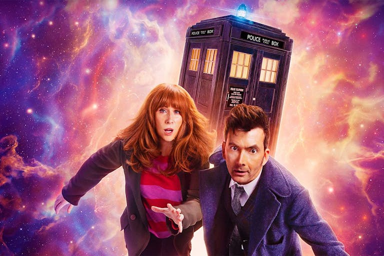 A red headed white woman and a dark haired white man in long blue coat, run away from cosmic galaxy activity with the TARDIS, a blue police box, in the background