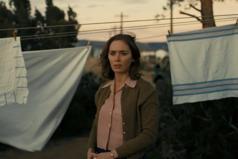 A white woman with short windswept brown hair looking concerned and thoughtful, with white sheets pegged on a clothes line behind her