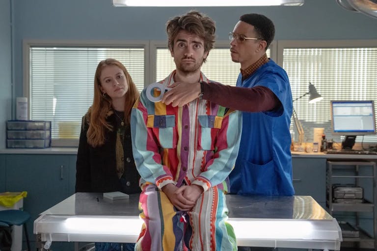 A young white man sits on a GPs table wearing a colourful jumpsuit with a Black male doctor scanning him with a devise, a young redheaded white woman stands in the background