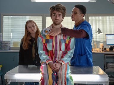 A young white man sits on a GPs table wearing a colourful jumpsuit with a Black male doctor scanning him with a devise, a young redheaded white woman stands in the background