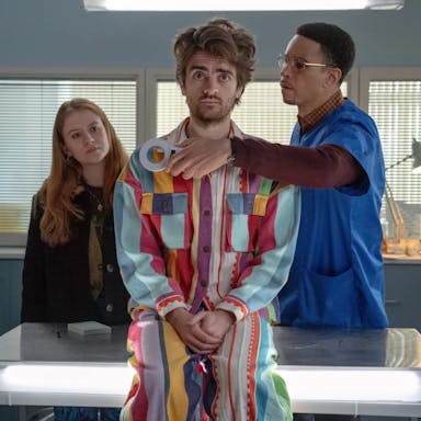 A young white man sits on a GPs table wearing a colourful jumpsuit with a Black male doctor scanning him with a devise, a young redheaded white woman stands in the background