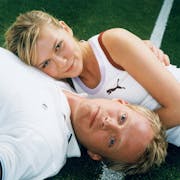 A blonde woman laying her head on a blond man's chest as he lays the opposite way to her, both in tennis clothing