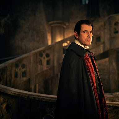 A seemingly middle aged white man with pale skin in a dark gothic setting wearing a black and red cape