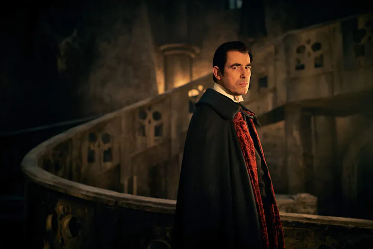 A seemingly middle aged white man with pale skin in a dark gothic setting wearing a black and red cape
