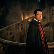 A seemingly middle aged white man with pale skin in a dark gothic setting wearing a black and red cape