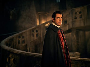 A seemingly middle aged white man with pale skin in a dark gothic setting wearing a black and red cape