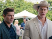 Two white men one in a dark casual suit, the other in a white suit and white cowboy hat, at a garden party