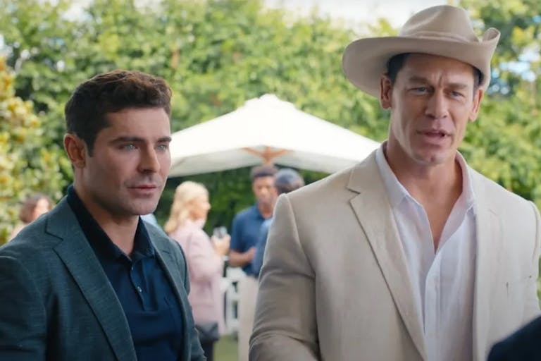 Two white men one in a dark casual suit, the other in a white suit and white cowboy hat, at a garden party