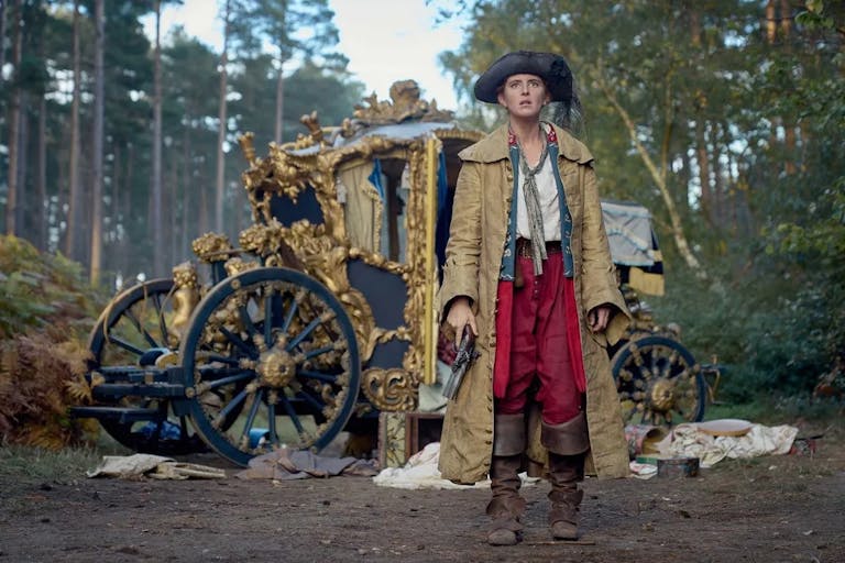 A young white woman in 18th Century pirate style clothing holding a pistol with an ornate broken down carriage behind her