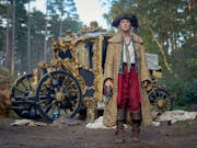 A young white woman in 18th Century pirate style clothing holding a pistol with an ornate broken down carriage behind her