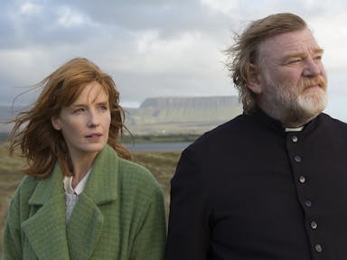 A woman and a man dressed as a priest stand in a windy Irish landscape.